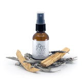 Amber spray bottle surrounded by white sage leaves and palo santo sticks. Smudge spray with essential oil blend infused with reiki by soul ritual to clear you energetic body and aura and clear your space of negativity